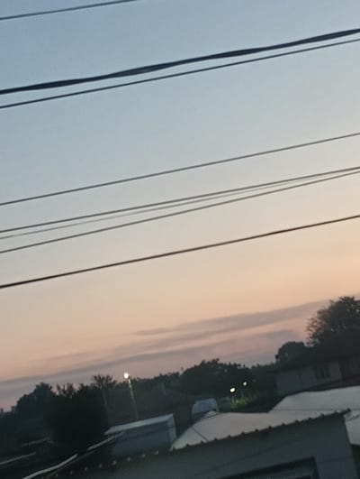 The image depicts an outdoor scene at either dawn or dusk, characterized by a gradient sky transitioning from light blue-grey at the top to soft peach and pink tones near the horizon. Several dark, thin utility wires traverse the upper half of the frame diagonally from the upper left to the lower right. Faint, elongated cloud formations are visible within the pink-hued sky. Below the horizon, a dark, indistinct band of trees and foliage is present. In the mid-ground, two small, bright, circular artificial light sources are visible against the dark foliage. The lower portion of the image reveals parts of multiple structures, including rooftops with grey tones, one appearing corrugated. The scene is located in the city of Straldzha, Bulgaria. No individuals or discernible actions are present.