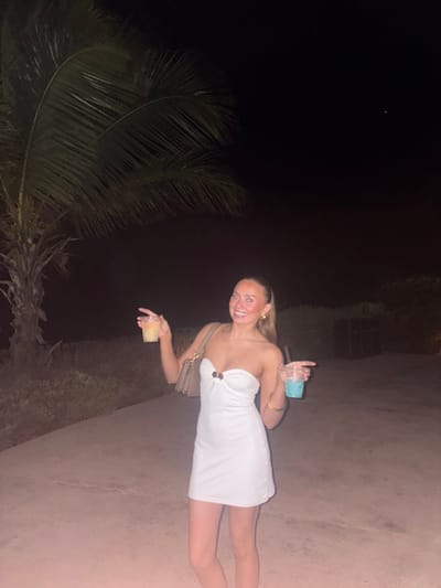 A woman in a white dress stands on a dimly lit path with a palm tree to her left. She holds a drink in each hand and smiles at the camera. The backdrop is a dark night sky. The woman's dress is short and fitted, and she has a small purse hanging from her shoulder. The path is made of concrete or stone. The palm tree is tall and has long, green fronds.
