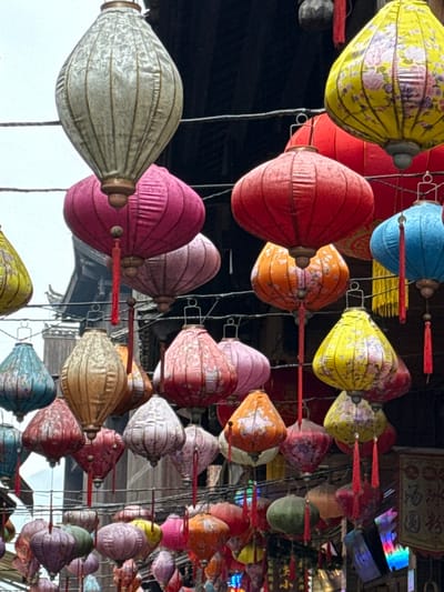 The image depicts a dense overhead canopy of numerous traditional Chinese lanterns suspended across what appears to be a street or pathway in Pengshui Miao and Tujia Autonomous County, China. The lanterns exhibit a variety of shapes, including elongated ovals, rounded spheres, flat-bottomed pear-like designs, and hexagonal forms. Their colors span a wide spectrum, prominently featuring magenta, red, yellow, orange, blue, pink, light green, gold/tan, and white/cream, with many adorned by intricate floral or pattern designs. Numerous lanterns feature hanging tassels, predominantly red. They are suspended by dark wires or ropes crisscrossing the frame. In the background, sections of traditional Chinese architecture are visible, characterized by dark wooden structures, tiled roofs, and some lighter-colored building facades. An overcast or hazy light grey sky is visible in the upper left portion of the frame. A partial sign with Chinese characters is visible in the lower right, suggesting a commercial or public thoroughfare. No individuals are visible.