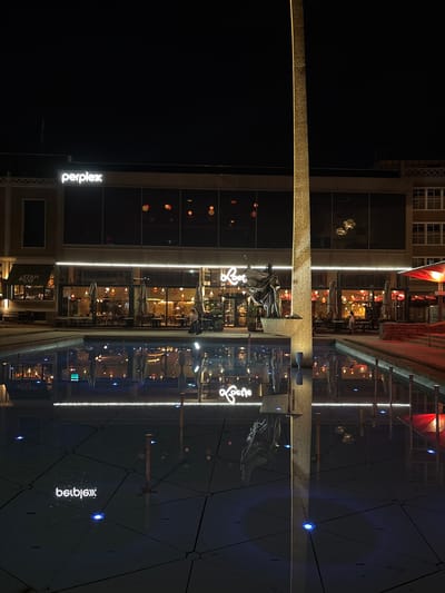 The image captures a nighttime view of the city of Arnhem, Netherlands, specifically focusing on the area surrounding the Plein. In the background, a multi-story building is illuminated, with a prominent neon sign spelling "perplex" on its upper facade. Below this, a row of businesses with large glass windows lines the street, revealing restaurant interiors with tables, chairs, and ambient lighting. The reflection of these establishments is visible on the wet surface of the Plein, a public square featuring a shallow water feature.
The Plein itself is paved with dark tiles arranged in a grid pattern, with several small blue lights embedded in the tiles, creating a subtle glow. The water surface of the fountain acts as a mirror, reflecting the building's lights, the "perplex" sign (appearing backward as "xelprep" in the reflection), and the illuminated signage of the businesses. A tall, obelisk-like monument, illuminated with a golden light from its base, stands prominently in the center-right of the frame, casting a strong reflection in the water.
In the foreground, the water feature is shallow, with distinct grid lines visible through the water, suggesting a tiled bottom. Several metal posts rise from the water's edge. The overall atmosphere is one of urban nightlife, with the interplay of artificial lights and their reflections creating a dynamic visual scene. A few people are visible in the reflections within the restaurant windows and near the entrance of one of the establishments.