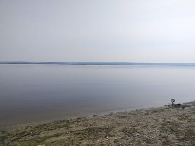 A serene, wide view of a calm, grey lake stretches towards a hazy, blue horizon under an overcast sky. The foreground features a gently sloping, pebbly shore with sparse green grass and a few scattered dry twigs. A dark, twisted object resembling a metal ring is partially visible on the shore near the right edge of the frame. The water's surface is smooth with subtle ripples, reflecting the muted tones of the sky. In the distance, a low, forested landmass is silhouetted against the sky. The scene depicts a natural landscape.