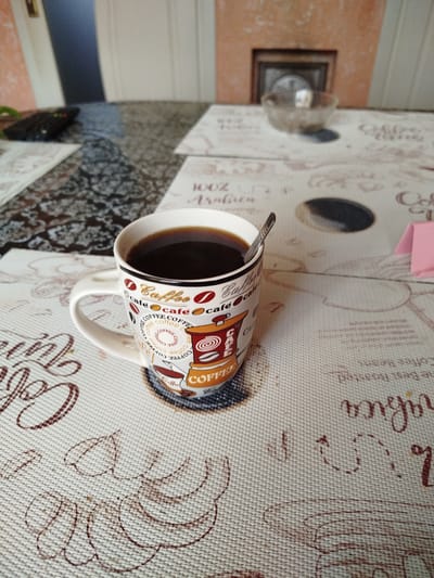 A high-angle medium shot captures a mug of coffee with a spoon resting inside on a textured, light-colored tablecloth featuring coffee-themed graphics and text. The mug is white with a colorful design depicting coffee beans, cups, and a coffee grinder. The coffee inside is dark brown and fills the mug nearly to the brim. To the left of the mug, part of a black remote control is visible on a darker, patterned surface. In the background, a blurred wall with an orange-toned section and what appears to be a decorative frame is visible. The scene is set indoors, likely on a table. No people are present in the image. The location of Pănășești, Moldova is not depicted in this image.