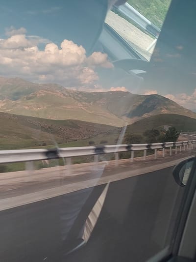The image captures a vast mountainous landscape viewed from inside a moving vehicle, likely a car. The primary scene features a paved road on the right side, extending into the distance, bordered by a multi-bar metal safety barrier. To the left of the road, a series of large, undulating mountains dominate the mid-ground and background. These mountains appear to be covered in sparse green and brown vegetation, suggesting a dry environment or a specific season. A small building or structure is faintly visible nestled among trees on one of the distant hills.
The sky, occupying the upper left portion of the image, is a clear blue, adorned with large, fluffy white cumulus clouds. The time of day is daytime, with ample natural light illuminating the scene, casting soft shadows on the mountains and clouds.
In the immediate foreground, elements of the car from which the photo was taken are visible, including a portion of a side mirror and the window frame in the upper right. A prominent reflection on the car's window in the lower left shows what appears to be another section of road and green grass, indicating the perspective from within the vehicle. The slight blur on the road barrier hints at the vehicle's movement.
The setting evokes a journey through a rural, mountainous region. Based on the provided context, the scene is located in or near Yelpin, Armenia. No people or visible text are present in the image.