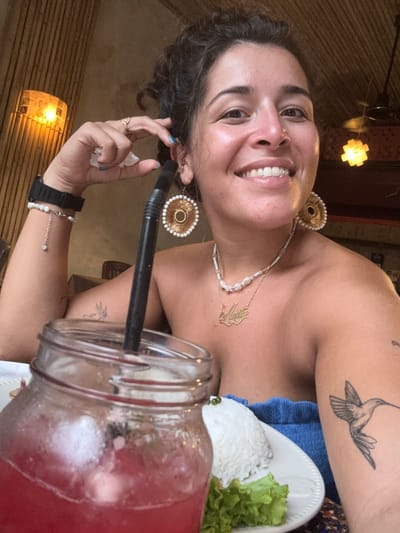 A woman with dark, curly hair pulled back sits at a table with a drink in a mason jar. She wears large, circular earrings with gold accents and white beads, a pearl necklace, and a gold nameplate necklace. A black watch is on her left wrist, and a bracelet with a small charm hangs from it. She has a hummingbird tattoo on her right arm. In front of her is a white plate with a mound of white rice and a leaf of lettuce. The background appears to be a restaurant with bamboo walls and warm lighting. The woman is smiling and holding a piece of tissue near her face. The scene is likely in Kampot, Cambodia.