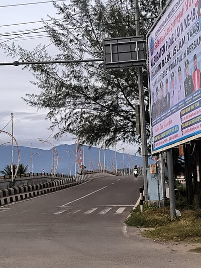 The image captures a street scene in Meureudu, Indonesia, featuring a road leading over a bridge, a billboard, and natural elements. The road surface is asphalt with white lane markings and a pedestrian crossing marked with white stripes. A motorcycle with a rider is visible on the road, heading away from the camera.

To the right of the road, a large billboard stands affixed to a pole. The billboard displays text in Indonesian, including "FORKOPIMDA KABUPATEN PIPIE JAYA KE-19," "TAHUN BARU ISLAM 1448," and "BANGKIT BERSYARIAT, PULIH BERSAM." Below this text are images of several men in formal attire, some wearing military uniforms.

Adjacent to the billboard, a utility box is mounted on a pole.

The bridge, spanning across what appears to be a body of water, has decorative metal arches and is lined with black and white striped barriers. Small Indonesian flags are visible on the bridge.

In the background, mountains are visible under a cloudy sky. A large tree with sparse foliage is prominent in the upper left portion of the image, with overhead power lines crisscrossing through its branches. The environment appears to be semi-urban, with some vegetation growing along the roadside. The overall lighting suggests it is either early morning or late afternoon.