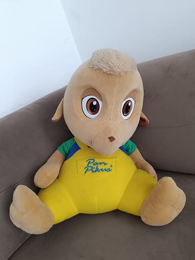 A plush toy, resembling a tan-colored animal with large brown eyes, is sitting on a brown sofa. The toy is wearing a yellow jumpsuit with blue straps over a green shirt. Blue embroidered text, "Pam Pikus," is visible on the front of the jumpsuit. The sofa is positioned against a white wall, suggesting an indoor domestic setting. The overall impression is a still life of a toy. The location in the city of Czekanów, Poland, is not directly discernible from the visual information provided in the image.