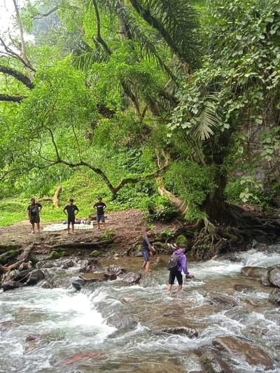 The image shows a small, rocky stream in the lush, green forest of Tawangmangu, Indonesia. Located at coordinates -7.655 latitude, 111.136 longitude, the scene reveals a group of six young men enjoying the natural setting. Two men are standing by the stream's edge while two others are wading in the water. One man is walking away from the camera along the stream bank. Two other men are sitting on a white blanket laid out on the dirt ground behind the others. The men are dressed casually, and the trees are thick and leafy. The water in the stream is shallow and flowing gently over smooth stones and rocks. The overall scene is peaceful and tranquil.
