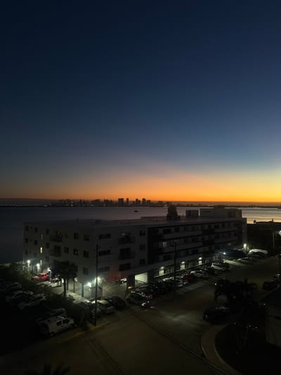 A cityscape at twilight reveals a building overlooking the water. The sky transitions from deep blue to orange and yellow hues. Silhouetted buildings line the horizon across the water. A parking lot beside the building is filled with cars, illuminated by streetlights. The scene captures the contrast between natural and urban environments at dusk.