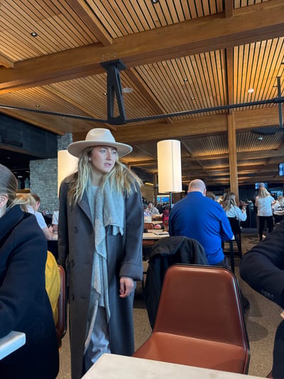 Inside a restaurant, a woman with blonde hair and a beige hat is wearing a gray coat and scarf. A ceiling lamp stands next to her, and wooden panels adorn the walls. Other people are seated at tables in the background, including a man in a blue shirt. The seating area appears busy and well-lit.