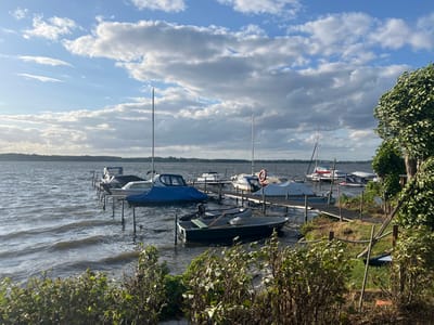 This image depicts a serene lake scene at a marina in Schwielowsee, Germany. Several boats, including sailboats and motorboats, are moored to a wooden dock. Some boats are covered with tarps, while others are open. The water is calm with gentle ripples, reflecting the cloudy sky above. Lush green bushes and trees line the shore in the foreground and to the right of the frame. The overall atmosphere is peaceful and tranquil, characteristic of a summer day at a German lakeside town.
