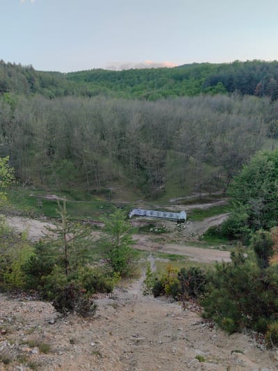 The scene depicts a view of a ravine or small valley in Slatina, Bulgaria. The vantage point is from a high, sandy or gravelly slope looking down towards a small stream or dry streambed. A white bridge or culvert spans the streambed. The landscape consists of mixed deciduous trees, some with leaves and some still bare, typical of early spring. A few small evergreen trees and bushes are visible in the foreground. In the background, a larger area of forest extends up the surrounding slopes. A crude wooden ladder is visible on the hillside to the left of the bridge. The sky is pale and slightly overcast. There are no people visible in the scene.