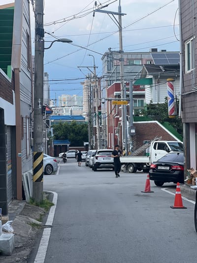 A street-level, medium-shot image captures a narrow urban alleyway in Gwangju, South Korea. The scene is dominated by buildings lining both sides of the street, featuring various architectural styles and materials, including brick, siding, and what appears to be metal panels. Overhead, a dense network of utility wires crisscrosses the sky, supported by concrete poles.

Several vehicles are parked along the right side of the street, including a white truck with a ladder on its bed, a black Kia sedan, and a white SUV. On the left side, a silver sedan is parked.

Two men are visible in the alley. One, wearing dark clothing and a cap, walks towards the viewer while looking at his phone. Another man, also in dark attire, walks away from the viewer on the right side of the road. A person on a bicycle is seen in the distance.

Orange traffic cones are placed on the road, and a barber pole stands vertically near the buildings on the right. Distant buildings with a noticeable presence of solar panels on their rooftops suggest a modern, albeit dense, urban environment. The sky is overcast with scattered clouds. The overall impression is one of a typical residential or commercial alleyway in a Korean city.