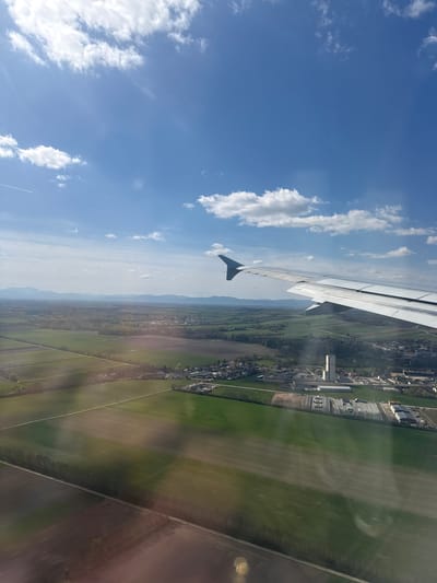 The image shows a view from an airplane window, with the plane's wing visible in the upper left. The landscape below consists of green fields and a town. The fields are divided by roads. A cluster of buildings, possibly an industrial area, is visible. In the distance are low-lying mountains. The sky is blue with scattered white clouds. The location is near the city of Enzersdorf an der Fischa, Austria, as suggested by the landscape and infrastructure.