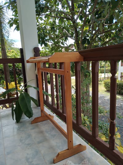 A wooden garment rack stands on a tiled balcony in front of a brown wooden railing, overlooking a garden area with lush green foliage and trees. A large potted plant with broad green leaves is positioned to the left of the garment rack. The balcony floor is made of light blue and white tiled squares. In the background, the scene transitions to a garden with gravel paths and various plants, suggesting an outdoor setting. The overall impression is one of a serene, tropical environment, likely in a residential area of Baan Mae Nam, Thailand.