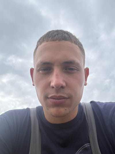 A close-up, low-angle selfie is taken of a young adult male with a close-cropped haircut. The man is looking directly at the camera, with his face occupying the majority of the frame. He is wearing a dark blue t-shirt with a gray strap crossing over his chest, possibly from a backpack. The background is a cloudy sky. The image provides no information about the location in Урыцкі сельскі Савет, Belarus.