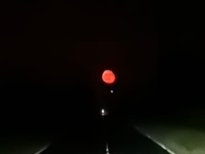 The image captures a deeply nocturnal scene on a road in Madrino, Bulgaria, viewed from the perspective of a vehicle looking forward into profound darkness. The most striking subject is a prominent, large, reddish-orange spherical object, likely the moon, centered in the upper portion of the frame. Its intense red hue suggests a "blood moon" event or strong atmospheric conditions affecting its appearance.

The road ahead is minimally visible, appearing as a dark, slightly reflective surface with faint white points of light intermittently along its centerline, possibly road markers or distant reflections. The surroundings on either side of the road are engulfed in deep blackness, indicating a rural or sparsely populated area completely devoid of artificial light. The time of day is clearly night. No specific weather conditions are discernible, though the moon's color hints at atmospheric factors. The scene evokes a sense of isolation and mystery under the expansive night sky, dominated by the unusual celestial display. There is no visible text in the image.