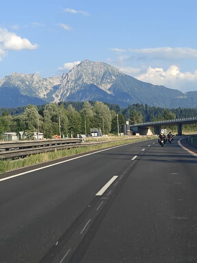 The image captures a view from a vehicle driving on a highway in Roßleithen, Austria, with the imposing Grimming mountain range in the background. The highway has two lanes visible, marked with white lines. Two motorcycles, ridden by individuals wearing helmets and protective gear, are traveling ahead on the highway. To the left of the highway, there is a grassy verge and a metal guardrail, beyond which are some buildings, possibly service areas or small structures. The background is dominated by lush green trees and the rugged, rocky slopes of the mountains, which are partially covered in vegetation and show distinct geological features. The sky is blue with scattered white clouds, indicating fair weather. A bridge or overpass is visible in the middle ground, spanning across the highway.