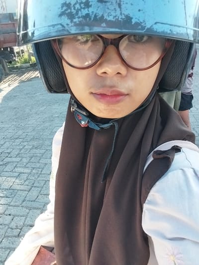 This is a portrait-style, self-taken photograph featuring a young woman wearing a brown hijab and large, round eyeglasses. She is also wearing a white shirt with what appears to be embroidered flowers on the collar and a brown outer garment, possibly a jacket or vest, that drapes over her. The woman has fair skin and her lips are a bright pink, suggesting makeup. A blue motorcycle helmet, which covers the left side of her face, is prominent in the foreground, with its visor reflecting some of the surroundings. The helmet strap is visible, secured with a buckle.

The background suggests an outdoor setting with a paved surface made of light-colored rectangular stones laid in a diamond pattern. In the further background, there are some green plants and a glimpse of vehicles, including what looks like a motorcycle and possibly a truck or car, indicating a semi-urban or roadside location. The lighting suggests it is daytime, likely late afternoon or early morning, given the warm, soft light. The woman's expression is neutral, and the close-up nature of the shot, combined with the helmet, gives a sense of a candid moment or a quick selfie. There is no visible text in the image. The orientation of the photo is landscape, with the subject appearing on the left.