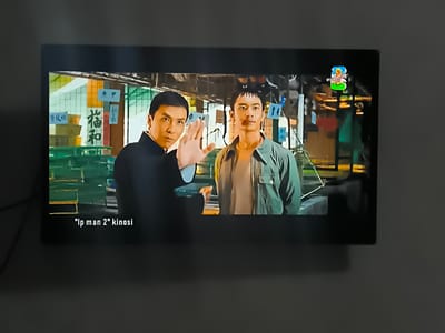 This is a media file depicting a scene from the movie "Ip Man 2". The image shows two men on a television screen. The man on the left, in a black martial arts uniform, is raising his right hand as if in a defensive or greeting gesture. He has a serious expression. To his right stands another man in a light green jacket over a tan shirt, looking directly at the camera with a neutral expression. Behind them, the setting appears to be an indoor market or a shop, with shelves stocked with goods and signs with Chinese characters visible. The lighting suggests it is daytime, but the indoor setting makes it difficult to determine the exact time of day or weather conditions. In the top right corner of the screen, a small logo is visible. At the bottom of the screen, text reads: "'Ip man 2' kinose". The overall mood is one of cinematic presentation, possibly a scene from a martial arts film. The location context provided is Kokand, Uzbekistan, suggesting this is how the movie is being presented or viewed there.