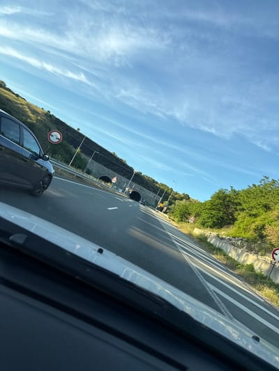 The image captures a perspective from within a vehicle traveling on a highway. The road ahead curves to the right, with multiple lanes marked by white lines. On the left side of the highway, a dark-colored car is visible in the adjacent lane. Beyond the car, a steep, grassy embankment rises, topped by a rock-faced wall and a tunnel entrance. A traffic sign with red and white circular markings and the text "50M" is positioned on the embankment. On the right side of the highway, a line of trees and vegetation borders the road, with a concrete barrier and more traffic signs visible. The sky is clear blue with scattered wispy clouds, illuminated by sunlight. The overall scene suggests a journey through a mountainous or hilly region, possibly a tunnel approach. Without specific landmarks or street signs, it is not possible to definitively confirm the location as Blatino, Bulgaria, though the scenery is consistent with such an area.