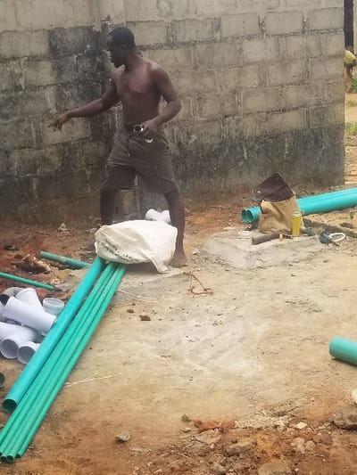 An adult male stands barefoot in the foreground of a construction site in Edu Town, Nigeria. He is shirtless, wearing dark cargo shorts, and holds a pair of sunglasses in his left hand. His right arm is extended, index finger pointing leftward, as he appears to be speaking. Behind him, a weathered concrete block wall forms the backdrop. On the ground to his left are stacked long teal PVC pipes and a pile of white PVC pipe fittings. A large white sack rests near his feet. To his right, a brown canvas bag and a yellow cylindrical object are visible on the ground. The terrain is composed of uneven dirt, soil, and patchy concrete. In the far right background, a second individual, wearing a yellow top, is partially visible, bent over.