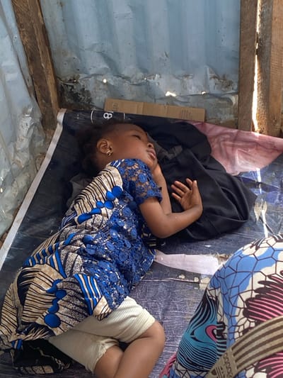 A young child is depicted lying on their left side, eyes closed, on a patterned mat within a makeshift structure in the city of 0801712591, Nigeria. The child wears a blue lace and patterned short-sleeved top over light-colored shorts or underwear, with a gold hoop earring visible in the right ear. Their left arm is bent, hand resting near the head, while the right arm is bent with the hand resting on a black fabric object. The mat, dark with white patterns and a visible number "8", is spread over an irregular surface. The background features weathered corrugated metal sheeting and rough wooden beams, suggesting a rudimentary shelter construction. A piece of cardboard is positioned against the corrugated metal. Natural light illuminates the scene from the right. In the lower right corner, a portion of another individual's leg, clad in brightly patterned fabric, is partially visible.