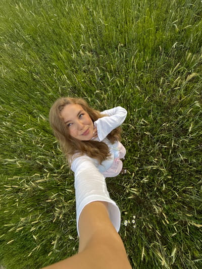 The image depicts a young woman taking a selfie in a field of tall green grass with some wheat stalks. She is wearing a white long-sleeved shirt and a light-colored dress with pink stripes. The woman has long, wavy brown hair and blue eyes. She is smiling and looking up at the camera. The field appears to be located in the village of Snovitsy, Russia, based on the provided context. The lighting suggests it is daytime, possibly late afternoon or early evening, with the sun casting a warm glow. There are a few small white flowers scattered in the grass near the woman's feet.