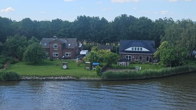 This is a medium-angle, eye-level drone shot capturing a waterfront property in Averlak, Germany, on a clear day with scattered clouds. The scene focuses on two brick houses situated on a grassy bank overlooking a body of water. The house on the left is older, with a mansard roof and visible dormer windows. A white caravan is parked in the yard. The house on the right is larger, with a dark, tiled mansard roof and a distinctive arched window. A small, octagonal garden house is located in front of this house. Both properties are surrounded by lush green lawns, mature trees, and tall reeds along the water's edge. Several children's play structures and outdoor seating areas are visible in the yards, suggesting a family-friendly environment. The water in the foreground is calm, reflecting the sky and surrounding vegetation. The overall impression is one of a serene, rural setting.