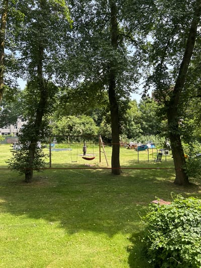 The image depicts a grassy yard in Ede, Netherlands, dominated by three large trees aligned in the foreground. A metal swing set is positioned between two of the trees. In the background, there's a glimpse of a building, possibly a residential structure. Further back, under the canopy of other trees, there is a collection of chairs, tables, and a trampoline visible. A patch of ivy or similar climbing plants is present on the right side of the image, growing on one of the trees. The overall scene suggests a public park, neighborhood green space, or private garden.