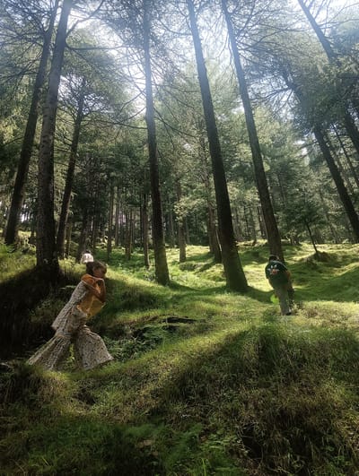 The image depicts a sun-dappled forest scene in Khakhnal, India, characterized by tall, slender trees with their branches reaching towards the sky, creating a dense canopy. Sunlight filters through the leaves, casting long shadows on the grassy, undulating terrain. In the foreground, a young girl with dark hair is shown from the chest up, wearing a patterned dress and a yellow scarf. She is looking downwards with her hand near her chin, possibly in thought or observation. Further back in the scene, a person wearing a dark green t-shirt with a logo and grey pants is walking away from the camera, ascending a slight incline. The environment is rich with green vegetation, including lush grass and ferns, indicating a natural, wooded area. The overall impression is one of a tranquil, outdoor excursion within a forest setting.