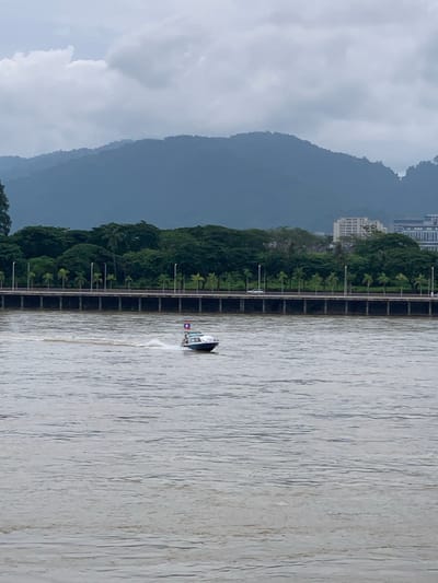 A small, white motorboat with a blue stripe is seen in the foreground, moving from left to right across a wide, murky river. The boat has a red and blue flag flying from its stern. In the middle ground, a multi-lane bridge spans the river, lined with streetlights and palm trees. Beyond the bridge, a dense line of green trees forms the middle ground. In the background, hazy blue mountains rise under a cloudy, overcast sky, with buildings visible on the right side of the frame. The overall impression is a somewhat muted and overcast day in a riverside city, likely Chiang Saen District, Thailand.