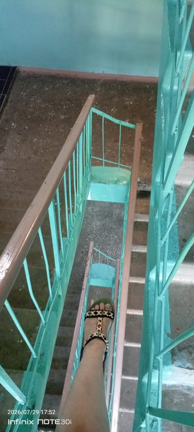 A first-person perspective shot shows a person's left leg and foot wearing a black sandal with gold and red embellishments. The sandal is adorned with a pattern of black straps and metallic accents, including what appear to be small round red beads or stones. The foot is positioned on a concrete staircase with teal-colored railings. The staircase appears to be part of an apartment building's interior, with worn concrete steps and a turquoise-painted landing visible in the background. The walls are painted a pale blue-green. The scene is lit by artificial light. The timestamp "2026/07/29 17:52" and the device model "Infinix NOTE 30i" are overlaid on the lower left of the image, indicating the date and time the photograph was taken, and the device used. The image does not provide enough information to determine the specific location within Langepas, Russia.