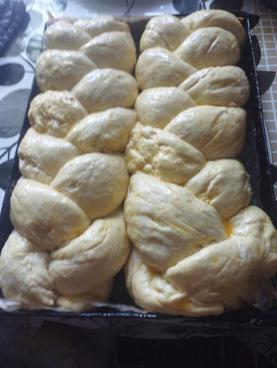 Two rows of braided challah dough, coated in an egg wash, are arranged in a black baking pan. The pan is placed on a table with a patterned tablecloth. The scene is set in Ciuciulea, Moldova.