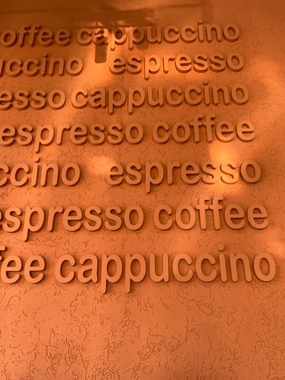 The image shows a textured orange wall with raised letters spelling out words related to coffee. The words are arranged in horizontal rows, with some letters partially obscured by shadows and the texture of the wall. From top to bottom, the visible words appear to be "coffee cappuccino," "ccino espresso," "esso cappuccino," "espresso coffee," "ccino espresso," "espresso coffee," and "ee cappuccino." The lighting suggests it is daytime, with sunlight casting shadows on the wall. There are no people or animals present in the image. The location is identified as Borisova Griva, Russia.