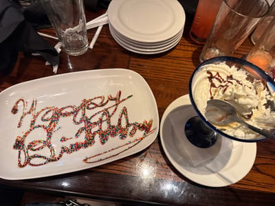 The image displays a tabletop scene, likely in a restaurant in Schaumburg, United States, captured after a birthday celebration. The central focus is a white, rectangular ceramic platter on which the words "Happy Birthday" are artfully written in chocolate syrup, heavily coated with vibrant, multi-colored sprinkles.
To the right of the birthday plate, a blue-rimmed glass dessert bowl contains the remnants of a sweet treat, primarily whipped cream with streaks of chocolate syrup, and a spoon resting inside, indicating it has been mostly eaten. This dessert bowl is placed on a small white saucer with a folded white napkin underneath.
The table is made of dark wood, showing reflections from the lighting. In the background, a dark cloth napkin is casually placed near an empty, clear drinking glass and some white paper items. A stack of five clean white round plates is neatly arranged further back on the left. On the right side of the background, several other empty or near-empty drinking glasses are visible, some with traces of a light pink or orange beverage. No people are present in the frame, but the arrangement suggests a recently concluded festive gathering.