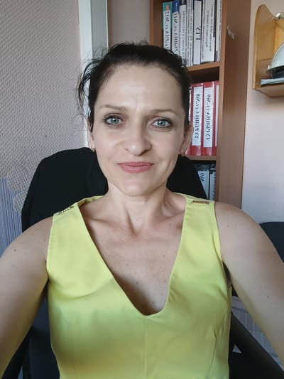 A woman with dark hair pulled back and light blue eyes is looking directly at the camera. She has on a yellow sleeveless dress with gold embellishments on the shoulders. The background includes a bookshelf filled with binders and a light-colored textured wall. This image was taken in Dnestrovsk, Moldova.