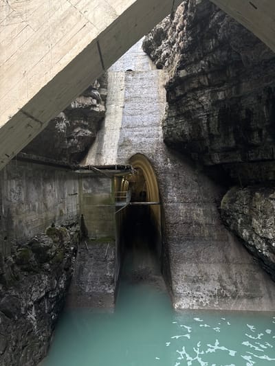 The image displays an engineered water control structure integrated into a natural rock gorge in Pellafol, France. A substantial, weathered concrete beam diagonally spans the upper left quadrant. Below this, a concrete channel contains light blue-green water in the foreground, flowing towards a dark, arched concrete tunnel entrance centrally positioned in the midground. Inside the tunnel entrance, an elevated concrete walkway with metal railings and illuminated by artificial lights is visible along the left interior wall. The water surface in the channel exhibits ripples and minor foam. Accumulations of dry branches and other debris are present at the waterline within the channel, immediately preceding the tunnel opening. The left side of the scene features a vertical concrete retaining wall partially merging with dark, moss-covered natural rock formations at its lower section. The right side is dominated by a dark, stratified natural rock face, into which the concrete arch structure is set. No individuals are present.