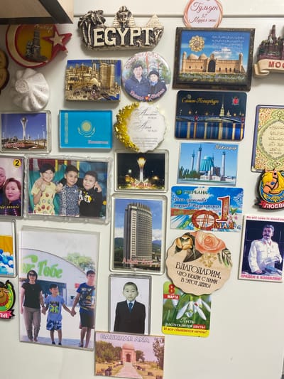 The image displays a collection of refrigerator magnets on a white surface, likely a refrigerator door. The magnets feature various landmarks, flags, and portraits, predominantly related to Kazakhstan and other countries. Specifically, there are magnets depicting: * **Landmarks:** * A prominent magnet shows a stylized Egyptian pyramid and sphinx with the word "EGYPTY" in large letters. * A magnet with a cityscape and minarets, labeled "Baku," likely representing Baku, Azerbaijan. * A magnet featuring the Baikonur Cosmodrome, with Russian text, likely related to space exploration. * A magnet showcasing the Astana Bayterek Tower, with the label "Астана" (Astana). Another similar magnet is also visible. * A magnet depicting a building with a domed roof, labeled "Тавбогар." * A magnet with a panoramic view of a city skyline and a bridge over water, labeled "Санкт-Петербург" (Saint Petersburg). * A magnet with a mosque and minarets, labeled "Тавбогар." * A magnet with a mosque and minarets, labeled "Tavbogar." * A magnet with a mosque, labeled "Bologdar." * A magnet with a building that appears to be the Central State Museum of Kazakhstan in Almaty, labeled "АЛМАТЫ" (Almaty). * A magnet with a tomb or mausoleum, labeled "GAUKHAR ANA." * **Flags:** * A magnet with the flag of Kazakhstan. * **Portraits and People:** * A magnet with a portrait of two young men and an adult male, with Cyrillic text below. * A magnet with a portrait of a man in a military-style uniform, with Cyrillic text "ПРИДЁМ К ИЗОБИЛИЮ!" (We will come to abundance!). * A magnet with a portrait of a woman and two boys, holding hands. The woman wears sunglasses and a black top, and the boys are in casual attire. The magnet has a green sign with Cyrillic text, potentially a store name. * A magnet with a portrait of a young boy in a suit and tie. * A magnet with a circular portrait of a man and a child, with Cyrillic text. * **Other Objects:** * A magnet shaped like a crescent moon with a star and a lighthouse. * A magnet shaped like a white, pleated object, possibly representing food. * A magnet with a decorative gold border and Cyrillic text. * A magnet with a stylized horse head. * A round magnet with a portrait of a man and a child and Cyrillic text. * A decorative wooden magnet with roses and Cyrillic text, "БЛАГОДАРИМ,