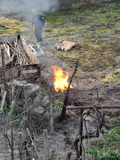 The image captures an outdoor scene in Krupnik, Bulgaria. One adult male, dressed in a dark jacket with an orange emblem on the upper left arm, dark pants, and light-colored shoes, is positioned in the upper left section of the frame. He is slightly bent forward, holding a dark, thin object directed towards a rudimentary wooden enclosure filled with dry sticks and debris. A prominent, active fire with bright orange and yellow flames is burning in the mid-ground, emitting a significant plume of white and gray smoke that rises and partially obscures the background. To the right of the male, on the ground, rests a light-colored, rectangular object with visible strapping or texture. The ground throughout the scene is primarily bare earth, interspersed with patches of sparse green vegetation. Numerous bare, gnarled branches and several upright posts, consistent with a vineyard or garden trellis system, are visible in the foreground and mid-ground.