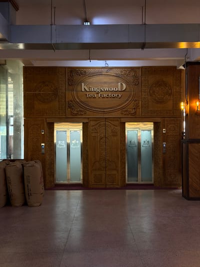 The image displays the interior of the Kingswood Tea Factory, located in Ramboda, Sri Lanka. The central focus is a large, intricately carved wooden wall featuring a prominent oval sign that reads "Kingswood Tea Factory" in raised lettering. This wooden paneling extends across the wall, exhibiting detailed circular and floral motifs.
Flanking a decorative, double-leaf wooden door in the center are two modern elevators with stainless steel doors and frosted glass panels. Each frosted glass panel bears the "Kingswood D" logo. Silver elevator call button panels, illuminated with blue arrows, are mounted on the wooden frame to the left of the left elevator and to the right of the right elevator. A dark red mat or carpet strip lies at the base of the elevator doors.
To the far left of the frame, partially obscured by incoming natural light from a large window or glass partition, are three large, light-colored fabric sacks, stacked vertically on the floor. These sacks show faint printed text, unreadable in detail. The structural elements of the building are visible through the glass on the left.
On the far right, a wooden pillar or wall section with a diagonal pattern is visible, adorned with two wall-mounted light fixtures featuring exposed filament bulbs. The ceiling above is light-colored, with exposed silver-colored rectangular ductwork supported by hanging rods. The floor is a light-colored terrazzo or polished concrete with darker aggregate. No people are present in the scene, and no actions or interactions are depicted.