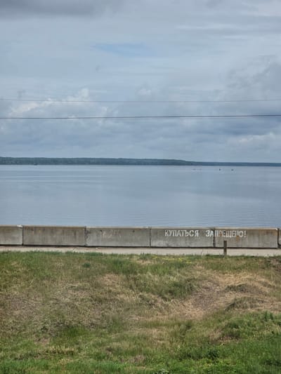 The image depicts a concrete barrier along the embankment of a wide body of water, likely a river or lake, under an overcast sky. The barrier, composed of several concrete blocks, is marked with white spray-painted Cyrillic text that reads "КУПАТЬСЯ ЗАПРЕЩЕНО!" which translates to "SWIMMING IS PROHIBITED!". In the distance, a treeline forms the horizon across the water. A few small boats or buoys are visible on the water's surface. In the foreground, a strip of dry and green grass is present. The scene appears to be located in Kozmodemyansk, Russia, based on the context provided.