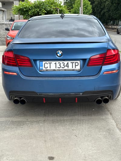 A blue BMW sedan is parked on a paved surface outdoors. The car's rear is facing the camera. The license plate reads "CT 1334 TP" and is bordered by the EU flag and the letters "BG". The car has quad exhaust pipes and a black diffuser with red accents. In the background, an orange car is partially visible, and behind that, there is a building with greenery. The scene appears to be in a residential area, possibly the city of Orizovo, Bulgaria, given the license plate format.