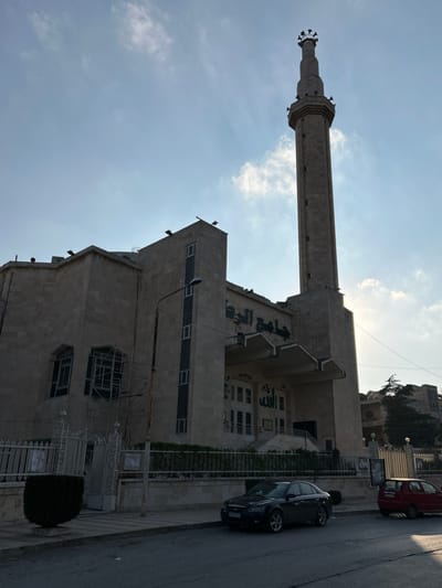 An outdoor, rotated full shot captures a modern mosque with a tall minaret under a bright, sunny sky, likely in Aleppo, Syria. The minaret, positioned on the left side of the building, is slender and cylindrical, constructed from light-colored stone, and features several decorative bands and what appear to be speaker mounts near its top. The sun, high in the sky, casts a strong backlight, creating some lens flare and highlighting the minaret's silhouette.
The mosque building itself is a multi-story structure made of light-colored stone, featuring numerous rectangular windows. Arabic calligraphy, in a dark contrasting color, adorns the facade; prominent among these characters, the word "مسجد" (Masjid, meaning Mosque) is visible, along with repetitions of "الله" (Allah) and other script. Scaffolding is visible on a lower section of the building, suggesting ongoing construction or renovation.
To the right of the mosque, a paved street runs parallel to the building. A dark-colored sedan is parked along the curb, and a portion of a red car is visible further down the street. A lamppost stands near the sedan. Green fencing and some sparse vegetation line the sidewalk next to the mosque. In the background, on a slightly elevated terrain, more residential buildings can be seen amidst greenery. The sky is largely clear with a few scattered clouds. The scene is static, capturing the architectural presence of the mosque and its urban surroundings during daytime.