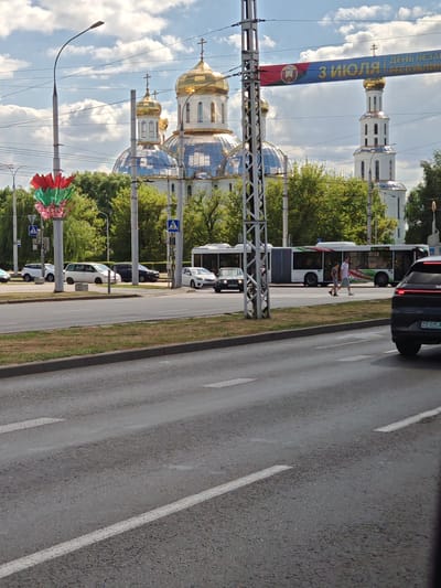 In the city of Alexandrovka, Russia, on a clear day with scattered clouds, a street scene unfolds. Dominating the background is a large, white Orthodox cathedral with multiple golden domes, reflecting the sky. To the right of the cathedral stands a tall, white bell tower, also topped with a golden dome. A banner with Cyrillic text and the Belarusian flag is suspended overhead, likely indicating a holiday or event.
In the mid-ground, a modern, articulated bus with a colorful design on its side is stopped, with passengers visible inside. Two individuals are walking across a pedestrian crossing in front of the bus. Several cars are parked and driving on the road, including a black SUV in the foreground. A street lamp with a decorative red, green, and white element is visible on the left. The environment includes a paved road, a grassy median, and a sidewalk lined with trees. The overall impression is of a bustling urban environment with significant cultural and religious landmarks.