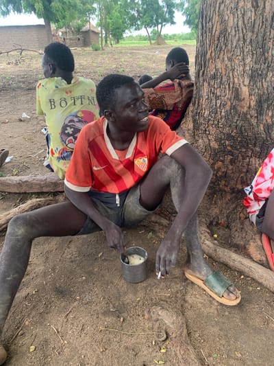 In Dull A, Nigeria, three individuals are seated outdoors near a large tree. The central figure, a dark-skinned male, wears an orange and red striped t-shirt and dark shorts. He is in a seated position with his legs crossed, holding a metal cup with a spoon. His right hand is near the cup, and his left hand holds a cigarette. His legs and arms appear to be dusty. To his left, another dark-skinned male wears a yellow t-shirt with the text "BOTTNI" printed on the back. To the right of the central figure, a third individual, whose face is not visible, wears a patterned fabric garment. The ground is composed of dirt and scattered debris, with tree roots visible. In the background, there are rudimentary structures that appear to be homes, and lush green trees. The lighting suggests it is daytime.