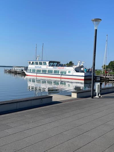 The image depicts a passenger ferry docked at a pier on a clear, sunny day. The ferry, named "Steinhude," is white with red stripes and has several windows along its side. Passengers are visible on the upper deck. The ferry is moored to a grey, wooden pier. In the background, there are sailboats with tall masts and trees. A modern-looking lamp post with a conical shade stands on the left side of the image. The water reflects the ferry and the sky. The scene appears to be a serene daytime setting at a harbor or lake. The text "Steinhude" and "meertouren.de" are visible on the side of the ferry.