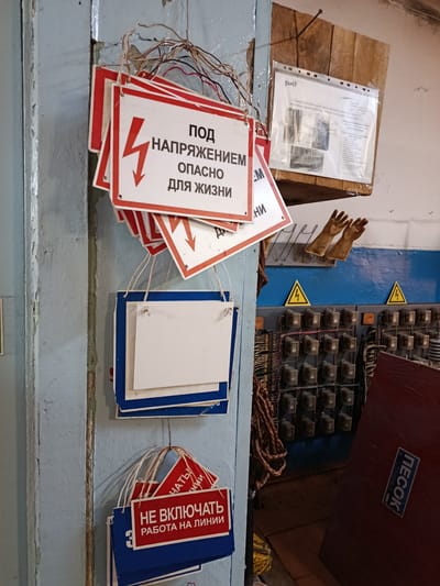 The image depicts an indoor scene, likely in a utility or industrial setting. Several red and white warning signs are hanging from wires on a light blue, weathered wall. One prominent sign reads "ПОД НАПРЯЖЕНИЕМ" (Under voltage) with a lightning bolt symbol. Below it, "ОПАСНО ДЛЯ ЖИЗНИ" (Danger to life) is visible. Other signs are partially obscured.

In the background, a wooden panel is mounted on the wall, displaying a document in a plastic sleeve. This document appears to be a technical instruction or diagram, with Russian text and illustrations. To the right of the panel, a pair of brown work gloves hangs on a rack.

Further to the right, a blue wall runs horizontally. Below it is a panel with numerous electrical switches or circuit breakers, with wires running into them. Two yellow triangular warning signs with lightning bolts are mounted on the blue wall.

In the foreground, a large dark red object with the white text "ПЕСОК" (Sand) in a blue rectangle is leaning against a wooden surface. The floor appears to be made of wooden planks. The overall environment suggests a place where electrical work or maintenance is conducted, with safety precautions prominently displayed. The mention of Urusha, Russia, indicates the geographical context of this scene.
