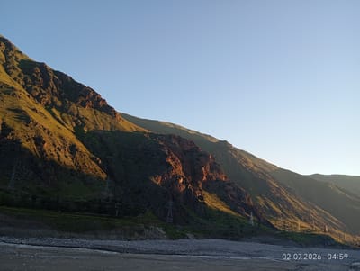 A wide shot captures a mountain range with steep, rocky slopes covered in sparse green vegetation. The sun, low in the sky, casts warm golden light on the upper portions of the mountains, creating stark shadows on the lower sections. A rocky riverbed or dry stream occupies the foreground, with a shallow stream of water flowing through it. Power line pylons are visible on the lower slopes of the mountains. The sky is a clear, pale blue. The timestamp "02.07.2026 04:59" is superimposed on the lower right corner of the image. The scene appears to be an undeveloped, natural landscape, likely in a mountainous region.