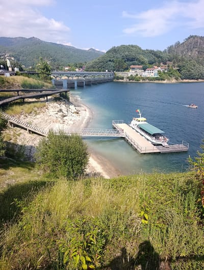 The image captures a scenic view of a reservoir with a modern pier and a white boat docked, located in the municipality of Terras de Bouro, Portugal. The reservoir water is a vibrant blue, reflecting the clear sky dotted with white clouds.
In the foreground, lush green vegetation and tall grasses cover a slope leading down towards the water's edge. A paved walkway with metal railings curves along the embankment, offering a path with elevated views of the surroundings. Below this walkway, a concrete staircase descends towards the sandy shore.
The pier, constructed from light-colored planks, extends into the water and is connected to the shore by a metal ramp. The white boat, identified by text on its side as "Rio Lima VI," is a passenger vessel with a green canopy covering its seating area. It displays the Portuguese flag at its stern. A smaller boat is visible in the distance, navigating the reservoir.
Across the water, the landscape is dominated by rolling hills covered in dense green forests. A large, arched bridge with multiple spans crosses the reservoir in the middle ground. On the far bank, a cluster of white buildings with red-tiled roofs is nestled among the trees. The overall impression is one of a tranquil natural setting enhanced by modern infrastructure for recreation and transport.