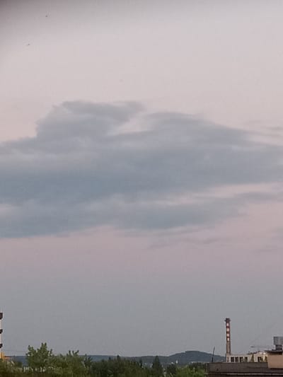The image depicts a cloudy sky over an urban landscape, likely in Kielce, Poland. The sky is a pale gray with soft pinkish hues, indicating either early morning or late evening. A large, amorphous cloud formation dominates the upper portion of the image. Below the clouds, a distant treeline is visible, suggesting a natural boundary or park area. In the middle ground, the rooftops of buildings can be seen, including a prominent industrial chimney with red and white stripes. The chimney is tall and narrow, topped with a section that appears to be under repair or disrepair, with exposed beams. Adjacent to the chimney are several low-rise industrial or commercial buildings. In the far distance, a hilly landscape is discernible, with a faint blueish tone. The overall impression is one of a somewhat overcast day in an industrial or semi-industrial part of the city. There are no people or animals visible in the frame.